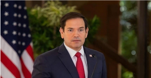 মাইক্রোফোন সামনে দাঁড়ানো মার্কো রুবিও টকশোতে বক্তব্য দিচ্ছেন; পটভূমিতে সংবাদ চিত্র ও বিশ্ব মানচিত্রের ঝলক।